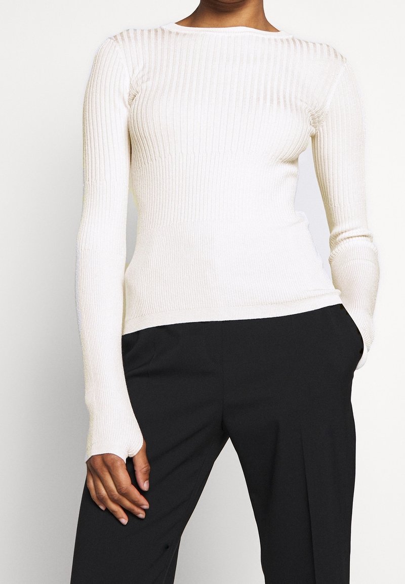 Femme portant un pull blanc côtelé ajusté à manches longues, associé à un pantalon noir ample, une main dans la poche, sur un fond uni.