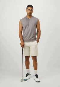 Férfi ujjatlan szürke trikóban és bézs sortban áll, egyik kezében zsebre dugja a kezét, a másikkal pedig golfütőt tart.