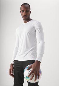 T-shirt de sport à manches longues blanc avec une texture, porté avec un pantalon noir, tout en tenant un ballon de football multicolore.