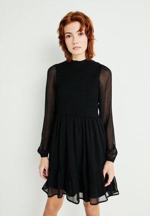 Robe noire avec un corsage smocké, des manches longues transparentes et une jupe à volants. Finition avec un ourlet à volants et un col rond.
