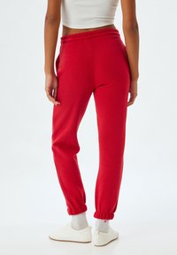 Røde sweatpants med snørelukning i taljen, afsluttede ankler, sidelommer og en blød tekstur, parret med hvide sneakers og lyse sokker.