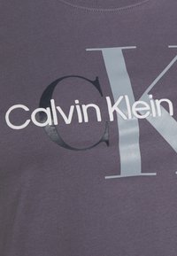 Camiseta morada con el texto "Calvin Klein" en blanco y negro, con una prominente letra "K" en gris. Textura de tela suave.