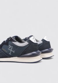 Zapatillas de deporte azul marino con parte superior de ante y nailon, que presentan acentos blancos, un diseño gráfico en el costado y una suela acolchada beige.