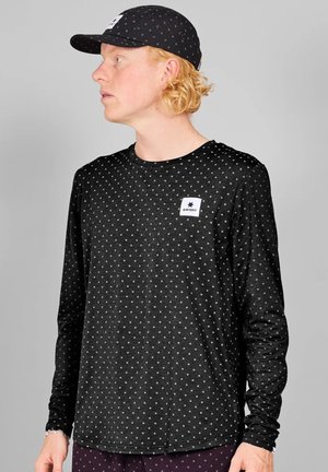 Mand med krøllet blondt hår iført en sort langærmet skjorte og kasket med små hvide polkaprikker, kigger mod venstre.