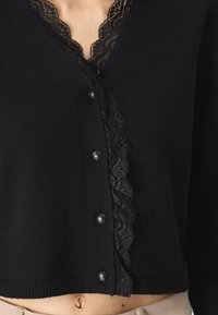 Melna kardigāna ar V veida kakla izgriezumu, ar melnas mežģīnes apdari priekšpusē un sešām melnām pogām. Mīksta adīta auduma tekstūra ar īsu hemu.