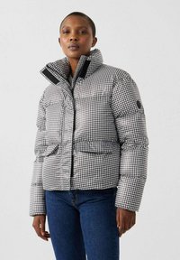 Chaqueta acolchada con patrón de pata de gallo en blanco y negro, con cuello alto, cierre de cremallera y dos bolsillos delanteros. Textura suave.