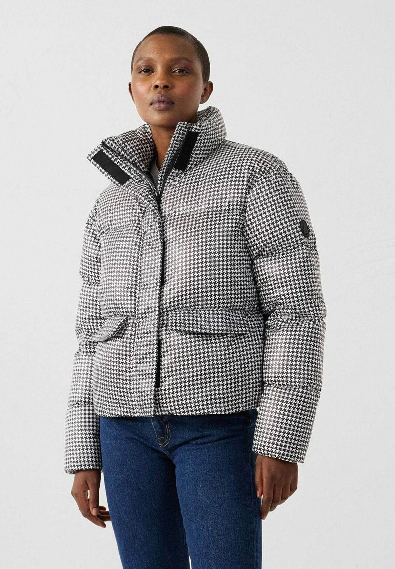 Chaqueta acolchada con patrón de pata de gallo en blanco y negro, con cuello alto, cierre de cremallera y dos bolsillos delanteros. Textura suave.