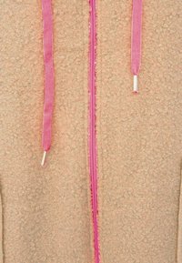 Beige textured fabric with a pink zipper and two pink drawstrings with silver aglets hanging down.