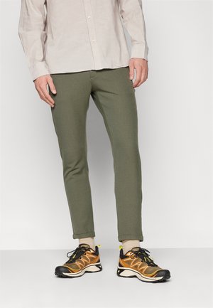 Olijfgroene tapered broek van een gestructureerde stof met omslag, gecombineerd met zwart-oranje sportschoenen en crèmekleurige sokken.
