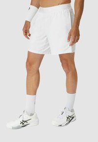 Atleta masculino vestido con pantalones cortos deportivos blancos, calcetines blancos, zapatos deportivos blancos y una cinta para la muñeca blanca, de pie contra un fondo liso.