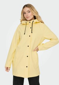 Vodoodporen rumen dežni plašč z dobro prileglo kapuco, opremljen s črnimi gumbi, vrvico za zategovanje in dvema stranskima žepoma. Gladke teksture, dizajn do kolen.