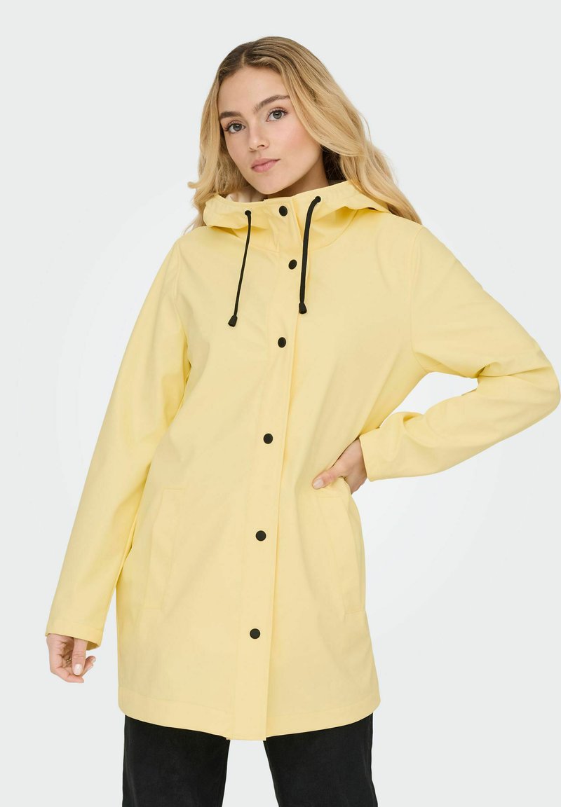 Vodoodporen rumen dežni plašč z dobro prileglo kapuco, opremljen s črnimi gumbi, vrvico za zategovanje in dvema stranskima žepoma. Gladke teksture, dizajn do kolen.