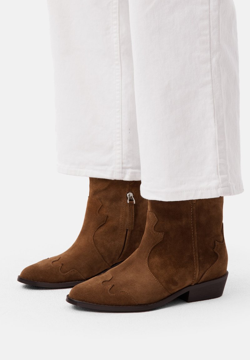 Stivali ankle in suede marrone con punta appuntita, ritagli decorativi, zip laterali e tacchi a blocco bassi, abbinati a pantaloni bianchi.