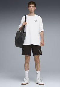 T-shirt blanc oversize avec un logo noir, short noir avec texte blanc, chaussettes blanches et baskets blanches. Sac à bandoulière noir. Fond gris.