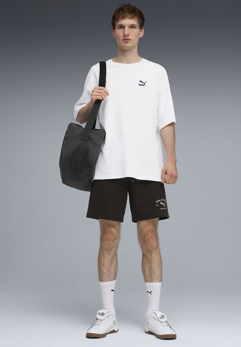 T-shirt blanc oversize avec un logo noir, short noir avec texte blanc, chaussettes blanches et baskets blanches. Sac à bandoulière noir. Fond gris.