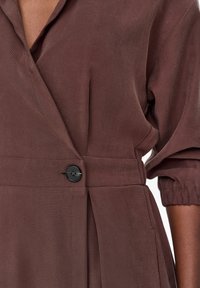 Brązowa sukienka kopertowa z teksturowanej tkaniny, szerokim kołnierzem, jednorzędowym czarnym zapięciem na guzik oraz elastycznymi mankietami z detalem plisowania.