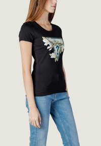Schwarzes T-Shirt mit kurzen Ärmeln, das ein florales Motiv in dreieckigem Design zeigt, akzentuiert mit blauen und grünen Details. Kombiniert mit blauen Jeans.
