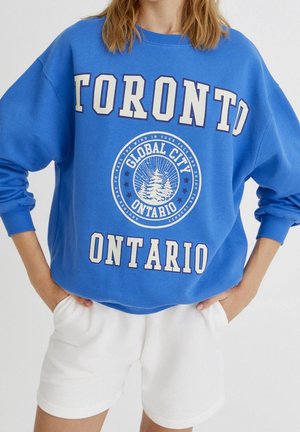 Mujer con sudadera azul con "Toronto Ontario" y un logo circular con árboles, combinada con pantalones cortos blancos, manos en los bolsillos.
