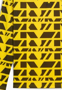 Gelbes Hemd mit einem geometrischen Muster aus braunen Formen, darunter Dreiecke und Rechtecke, die in verschiedenen Anordnungen über den Stoff verteilt sind.