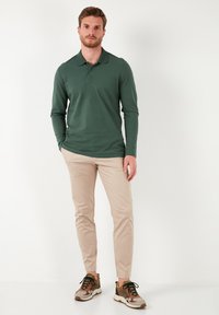 Camisa polo verde de manga larga combinada con pantalones slim-fit beige y zapatillas multicolores con acentos en marrón y amarillo.