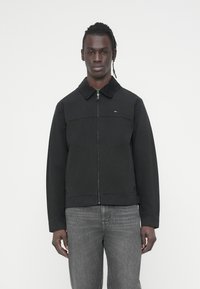 Μαύρο μπουφάν με φερμουάρ, με γιακά και μπροστινή επιφάνεια από κορντόρι, που διαθέτει μικρή λεπτομέρεια λογότυπου και ίσια γραμμή σχεδίασης. Φορεμένο με γκρι τζιν.