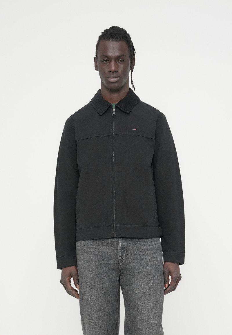 Μαύρο μπουφάν με φερμουάρ, με γιακά και μπροστινή επιφάνεια από κορντόρι, που διαθέτει μικρή λεπτομέρεια λογότυπου και ίσια γραμμή σχεδίασης. Φορεμένο με γκρι τζιν.