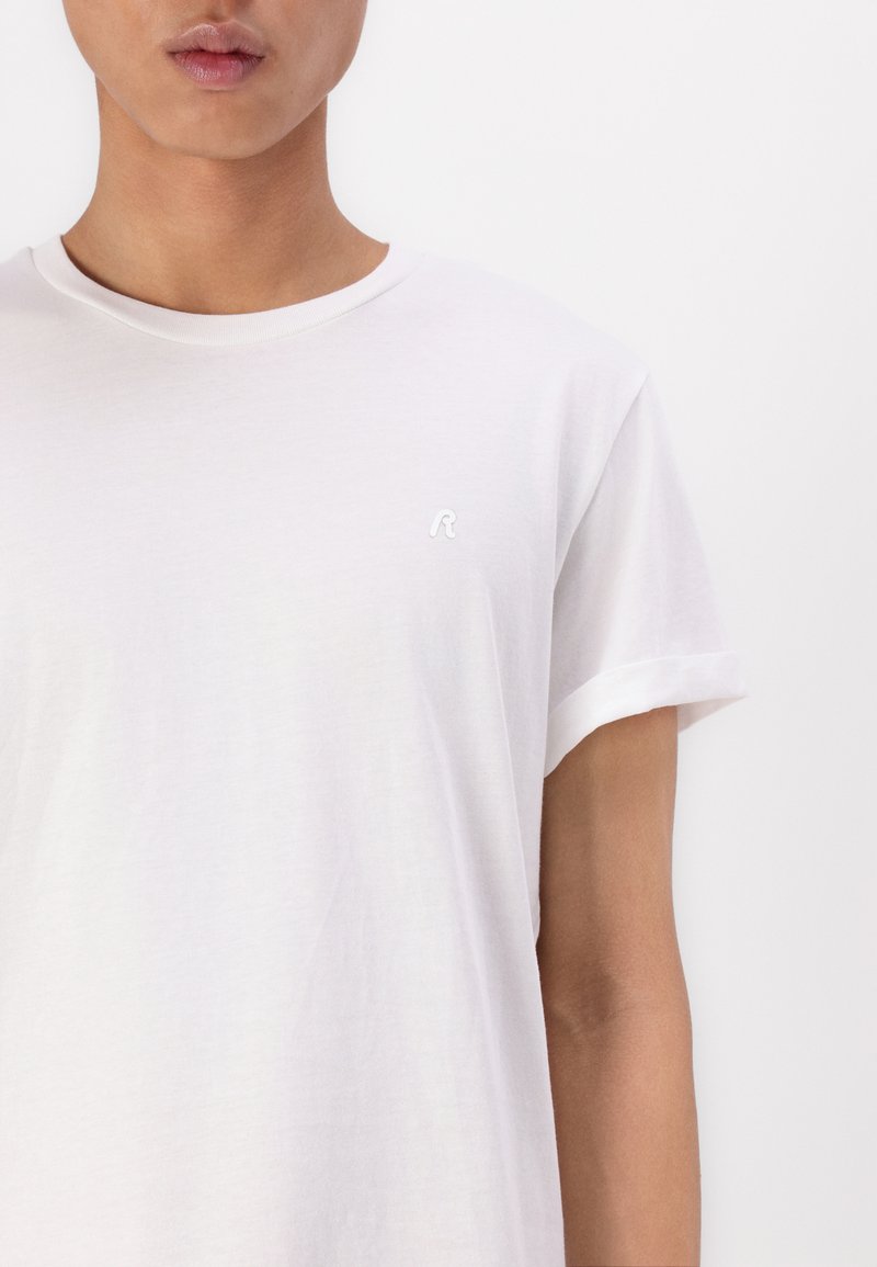 Weißes Baumwoll-T-Shirt mit hochgerollten Ärmeln, das ein kleines gesticktes Logo auf der Brust trägt. Glattes Material, lässiges Design.