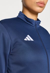 Αθλητικό μπουφάν σε ναυτικό μπλε με ψηλό γιακά, διαθέτει φερμουάρ στη μέση και λευκό λογότυπο με τρεις ρίγες στο αριστερό στήθος. Ομαλή υφή.