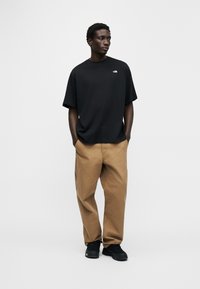 Άνδρας ντυμένος με μαύρο, φαρδύ t-shirt, καφέ παντελόνι φαρδύ στα πόδια και μαύρα αθλητικά παπούτσια, στέκεται με τα χέρια στις τσέπες, κοιτώντας προς τα πλάγια.