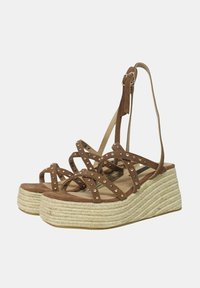 Sandali con zeppa in suede marrone, dotati di cinturini incrociati, con borchie metalliche e una piattaforma intrecciata simile al juta. I cinturini si attaccano alla caviglia.