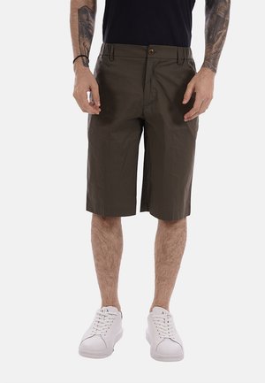 Uomo che indossa pantaloncini marroni fino al ginocchio, camicia nera, scarpe da ginnastica bianche, con braccia tatuate e mani rilassate ai lati.