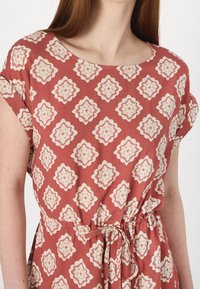 Robe couleur rouille avec un motif floral en crème. Manches courtes, col rond et ceinture à la taille pour cintrer. Texture de tissu douce.