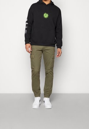 Felpa nera con grafica verde sul petto e maniche a motivi, abbinata a pantaloni cargo olive e scarpe da ginnastica bianche.