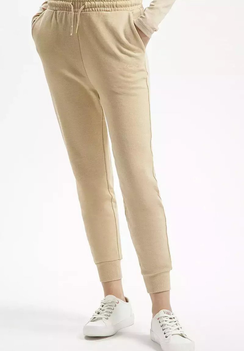 Beżowe dresy z teksturowanym wykończeniem, zwężanym krojem i elastycznym pasem. Zawierają boczne kieszenie i ściągacze przy kostkach.