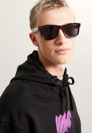 Gafas de sol con montura carey marrón y lentes oscuros, varillas grises con marca visible. Llevadas con una sudadera negra con capucha. Diseño simple y moderno.