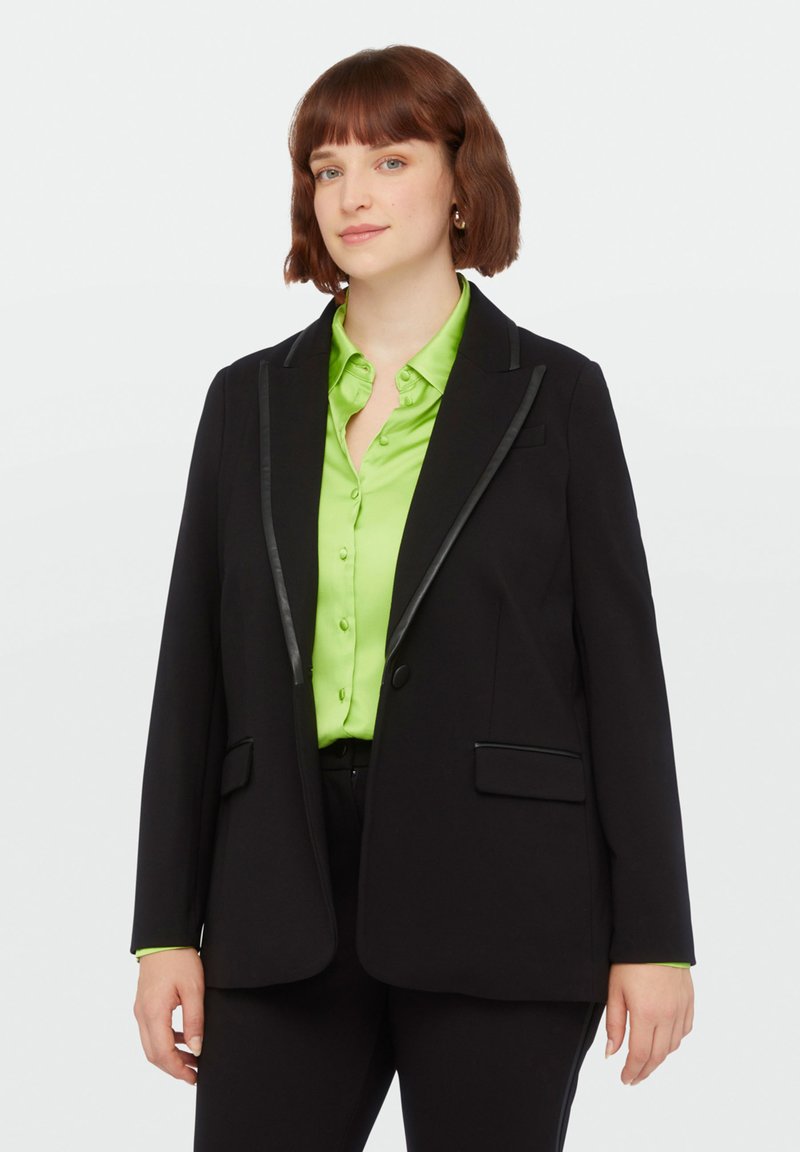 Blazer noir avec un revers noir contrastant, associé à une chemise à boutons vert vif. Comprend deux poches avant et une coupe décontractée.