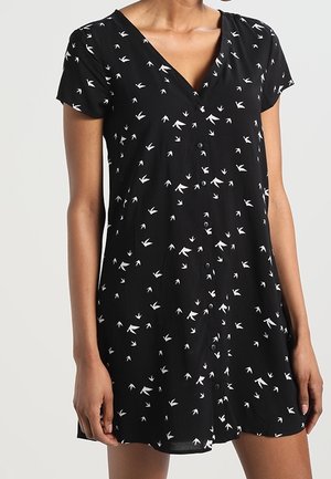 Schwarzes kurzärmeliges Hemdkleid mit V-Ausschnitt und kleinem Muster aus weißen Vogelfußabdrücken, getragen von einer Person.