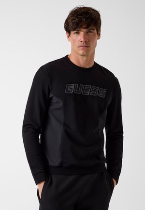 Młody mężczyzna o brązowych włosach, ubrany w czarną bluzę z długim rękawem z logo "GUESS", stojący z rękami w kieszeniach.