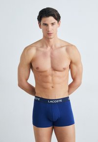 Oberkörperfreier Mann mit kurzen dunklen Haaren, der blaue Lacoste-Boxershorts trägt und mit den Händen hinter dem Rücken vor einem schlichten, hellen Hintergrund steht.