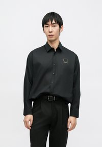 Crna košulja s dugim rukavima s diskretnim zlatnim vezenim uzorkom na lijevoj strani prsa, uparena s crnim hlačama i remenom.