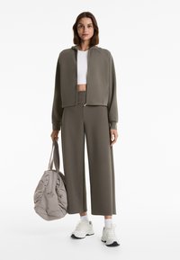 Olivgrüne, kurz geschnittene Kapuzenjacke mit Reißverschluss, weißes Crop-Top, dazu passende weit geschnittene Hose. Hält eine graue Tasche, trägt weiße Sneaker mit schwarzen Akzenten.