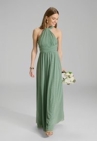 Robe longue à col halter en tissu vert doux avec une silhouette fluide ; comporte un corsage froncé et une texture lisse. Tient un bouquet.