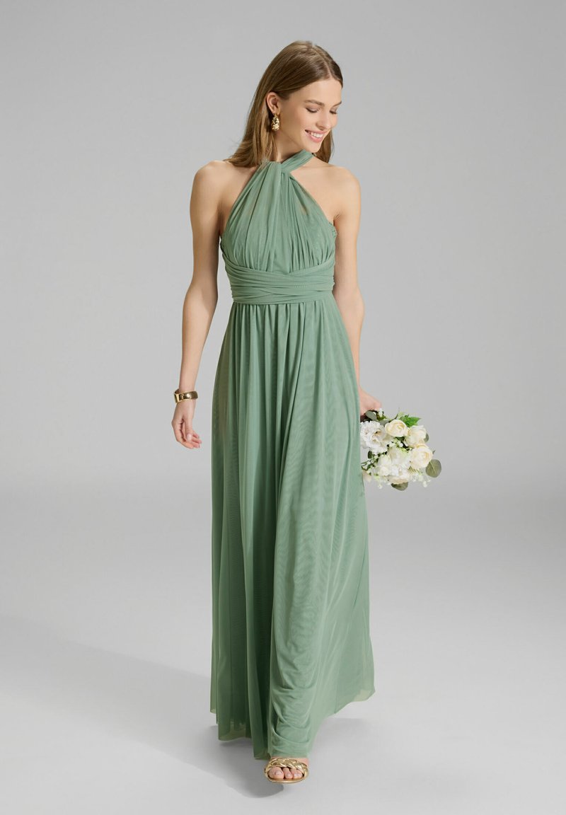 Robe longue à col halter en tissu vert doux avec une silhouette fluide ; comporte un corsage froncé et une texture lisse. Tient un bouquet.