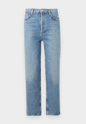 Kvinde med kort hår iført en hvid kortærmet ribbet top, højtsiddende cropped blå jeans og sorte sandaler med hæl, stående mod en ensfarvet baggrund.