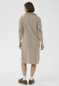 Donna vista da dietro che indossa un abito midi a coste beige con maniche lunghe, calze bianche e scarpe con zeppa marroni su uno sfondo bianco.