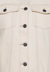 Cremefarvet denimjakke med sorte knapper, brystlommer og kontraststing. Struktureret design med en let tekstur.