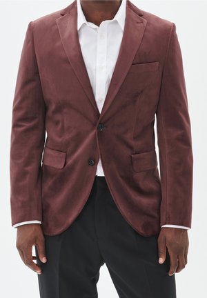 Hombre con una blazer de terciopelo burdeos sobre una camisa blanca de vestir y pantalones de vestir negros, de pie con los brazos relajados a los lados.