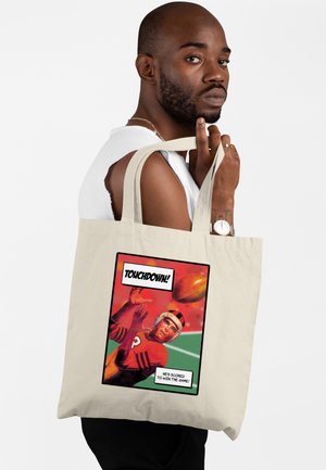 Muž držící béžovou tašku na rameno s komiksovým fotbalovým motivem a nápisem "TOUCHDOWN! Střelil vítězný bod!" na bílém pozadí.