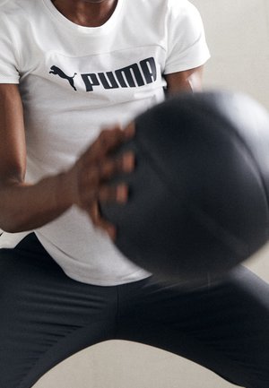 Personne portant un t-shirt blanc Puma et un pantalon noir tient et s'exerce avec un ballon médicinal noir sur un fond uni.