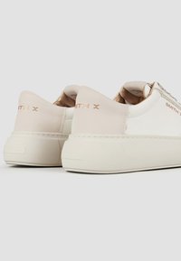 Sneaker bianchi in pelle con un morbido tallone beige, dettagli dorati, suola in gomma texture e marchio sul tallone. I dettagli di cucitura sono visibili.